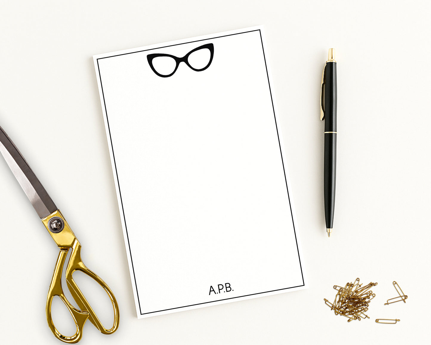 Custom notepad paper with a cat eye glasses design, personalized with monogram initials alongside a scissors, pen, and pins on a white background