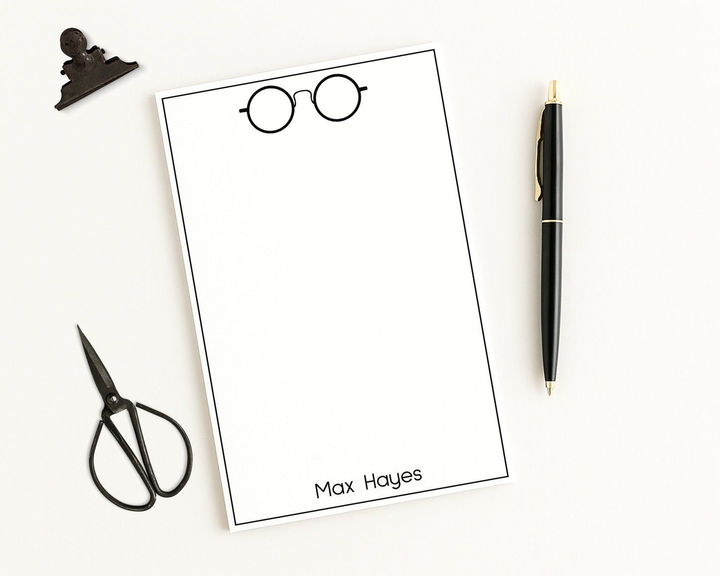 Personalized note pad paper with a name and glasses illustration, surrounded by stationery items on a white background