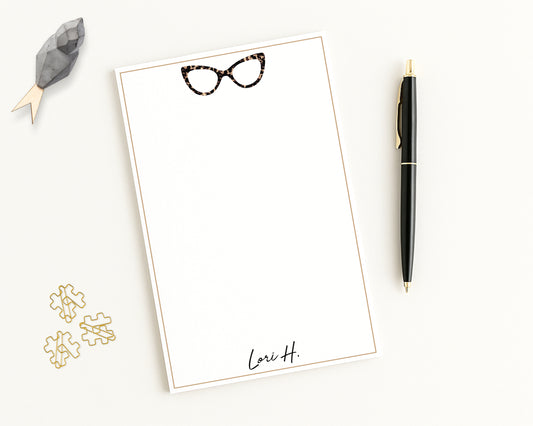 Personalized notepad features cat eye eyeglasses with custom monogram name, and beige border - shown with desk accessories.