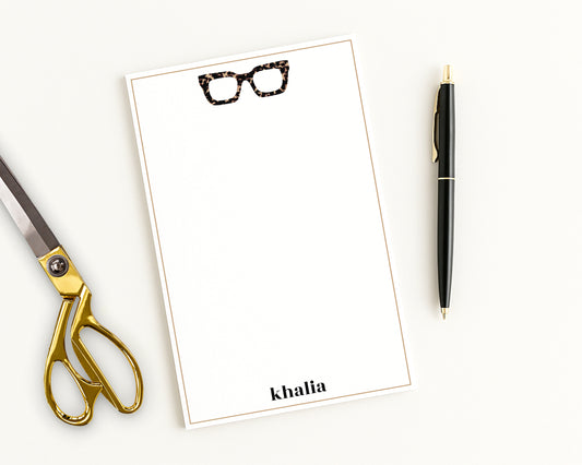 Personalized stationery notepad with chunky tortoiseshell eyeglasses icon and custom name, shown with desk accessories.