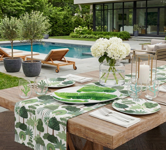 Outdoor dining setup by a pool with tropical tree print table runner and plates, banana leaf platter, coral motif glassware