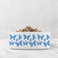 Hand drawn dancing butterflies in sky blue are printed on a white ceramic dog bowl.