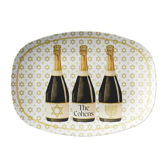 White oval serving platter features a graphic of three watercolor illustrated champagne bottles, each with a unique Hanukkah-inspired label:  one at the center is personalized with a name or phrase of your choice, one with a Star of David in shimmering gold glitter, and one featuring a glittery golden menorah. The entire tray is finished with an all-over Star of David print background in gold for an extra festive touch.