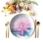 Dinner setting with a decorative plate featuring a pink water lily flower design, accompanied by gold cutlery and flowers on a white background.