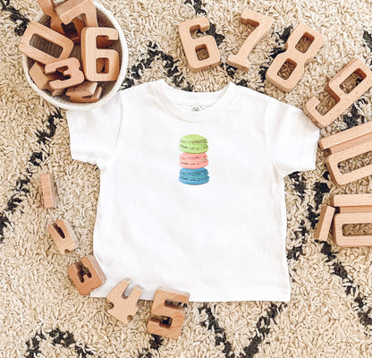 A white baby t-shirt with a stacked macaron cookie design in different colors placed on a floor with wooden blocks spelling out numbers.