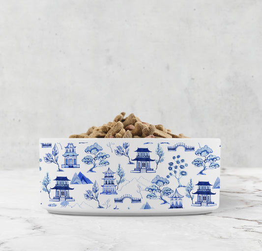Ceramic dog bowl with a blue and white chinoiserie pattern featuring pagodas, mountains and trees.