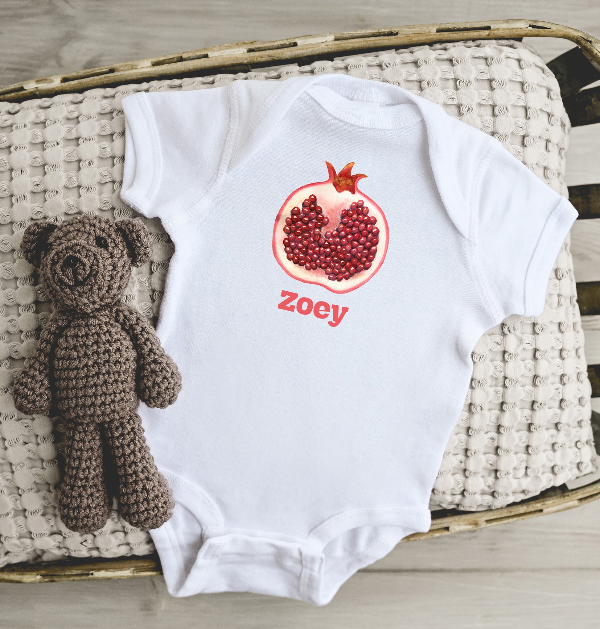 White baby onesie with a pomegranate design and 'zoey' text, accompanied by a brown crocheted teddy bear on a woven surface.