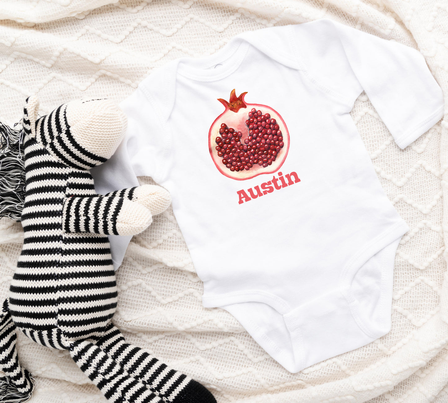 Baby onesie with pomegranate design and 'Austin' text, next to a striped stuffed toy on a textured surface.