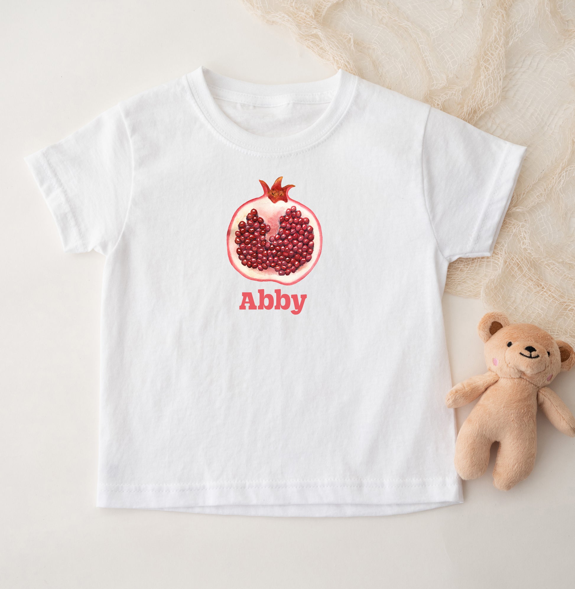 White t-shirt with a pomegranate design and 'Abby' text, next to a teddy bear on a light background
