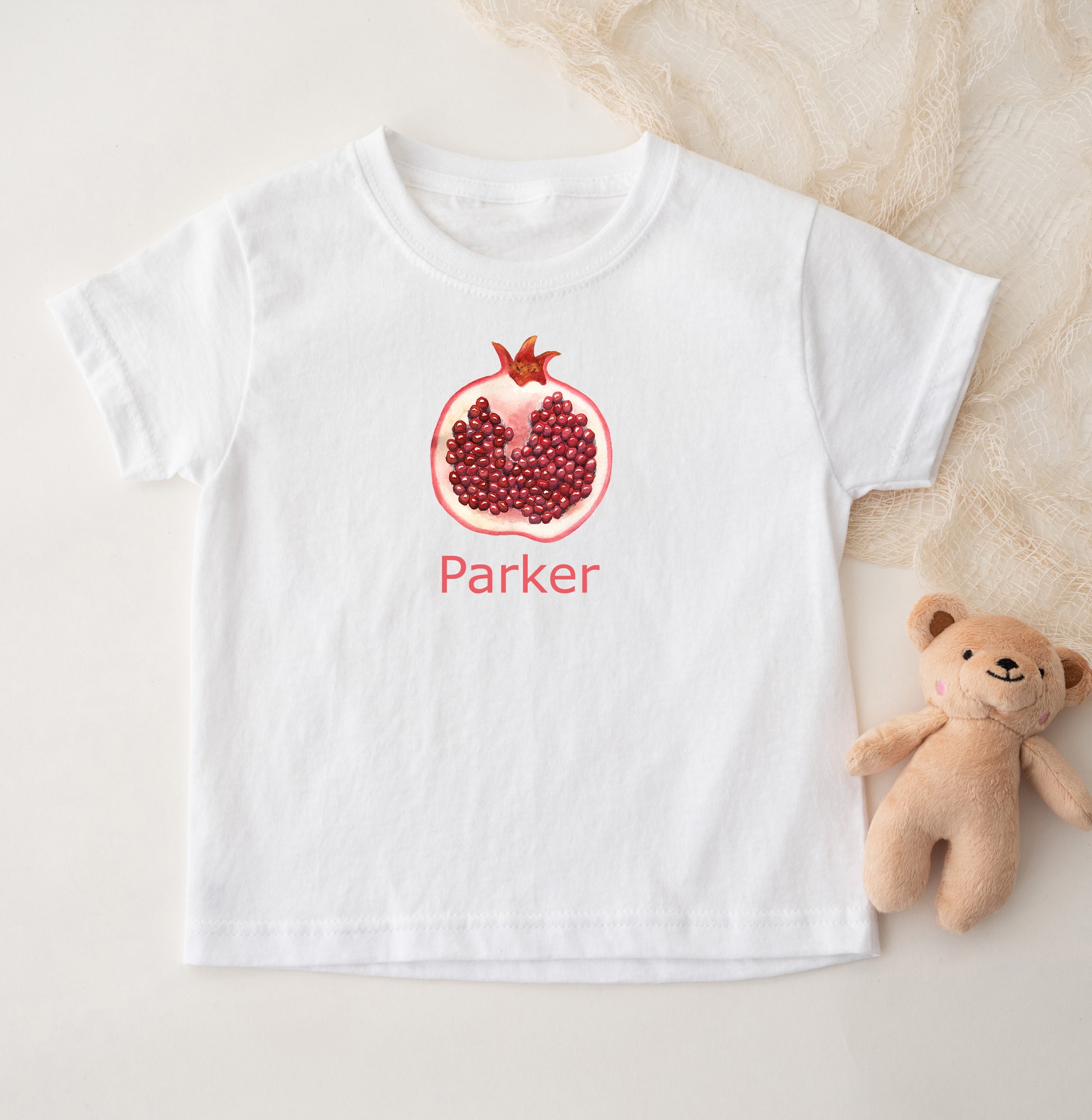 White t-shirt with a pomegranate design and 'Parker' text, accompanied by a teddy bear on a light background