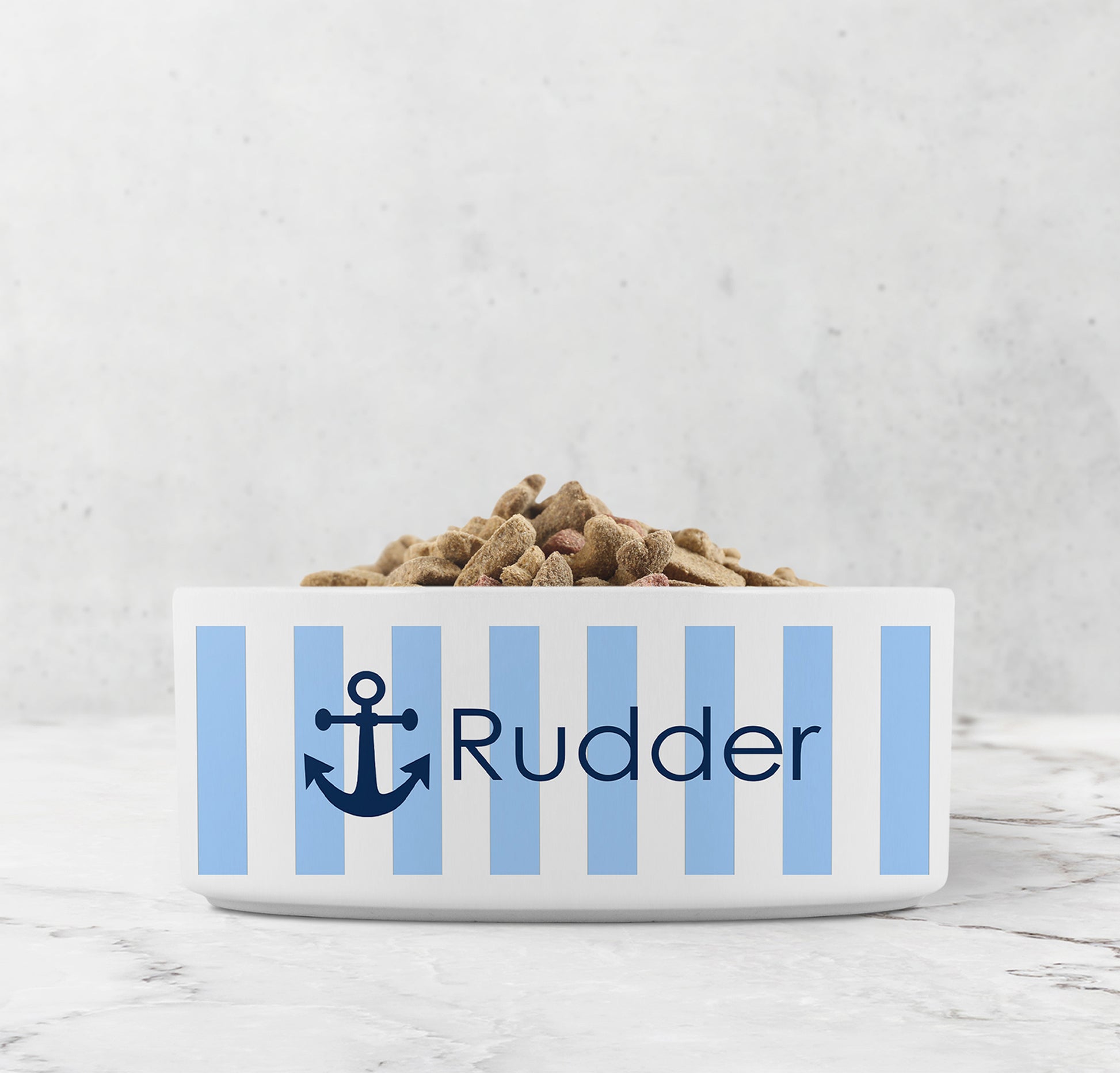 Dog or cat food bowl with blue and white striped pattern and navy blue anchor design with custom name on a marble surface.