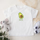 White t-shirt with avocado graphic and 'Maya' text, placed on a light surface with a plush toy and shoes.