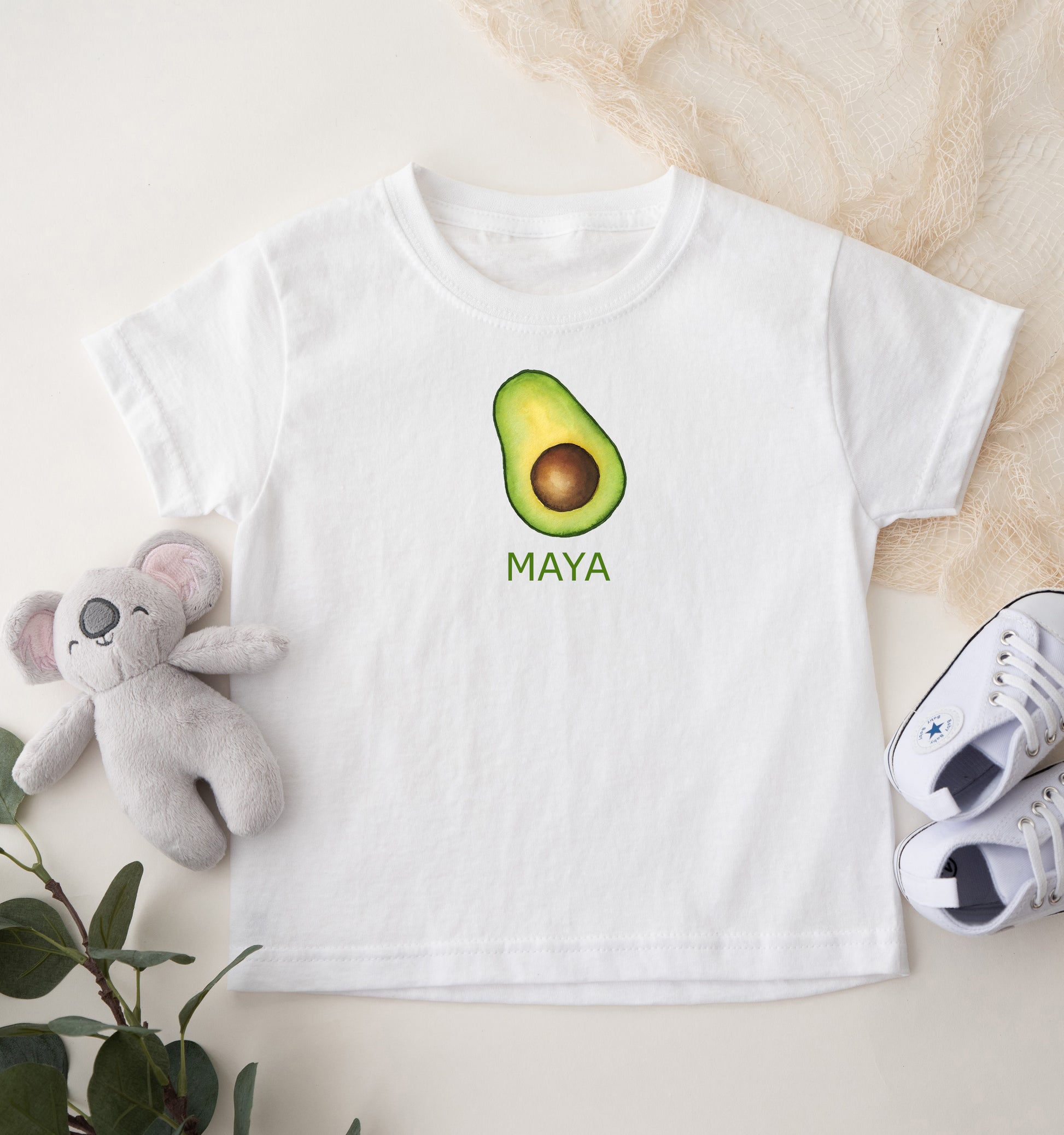 White t-shirt with avocado graphic and 'Maya' text, placed on a light surface with a plush toy and shoes.
