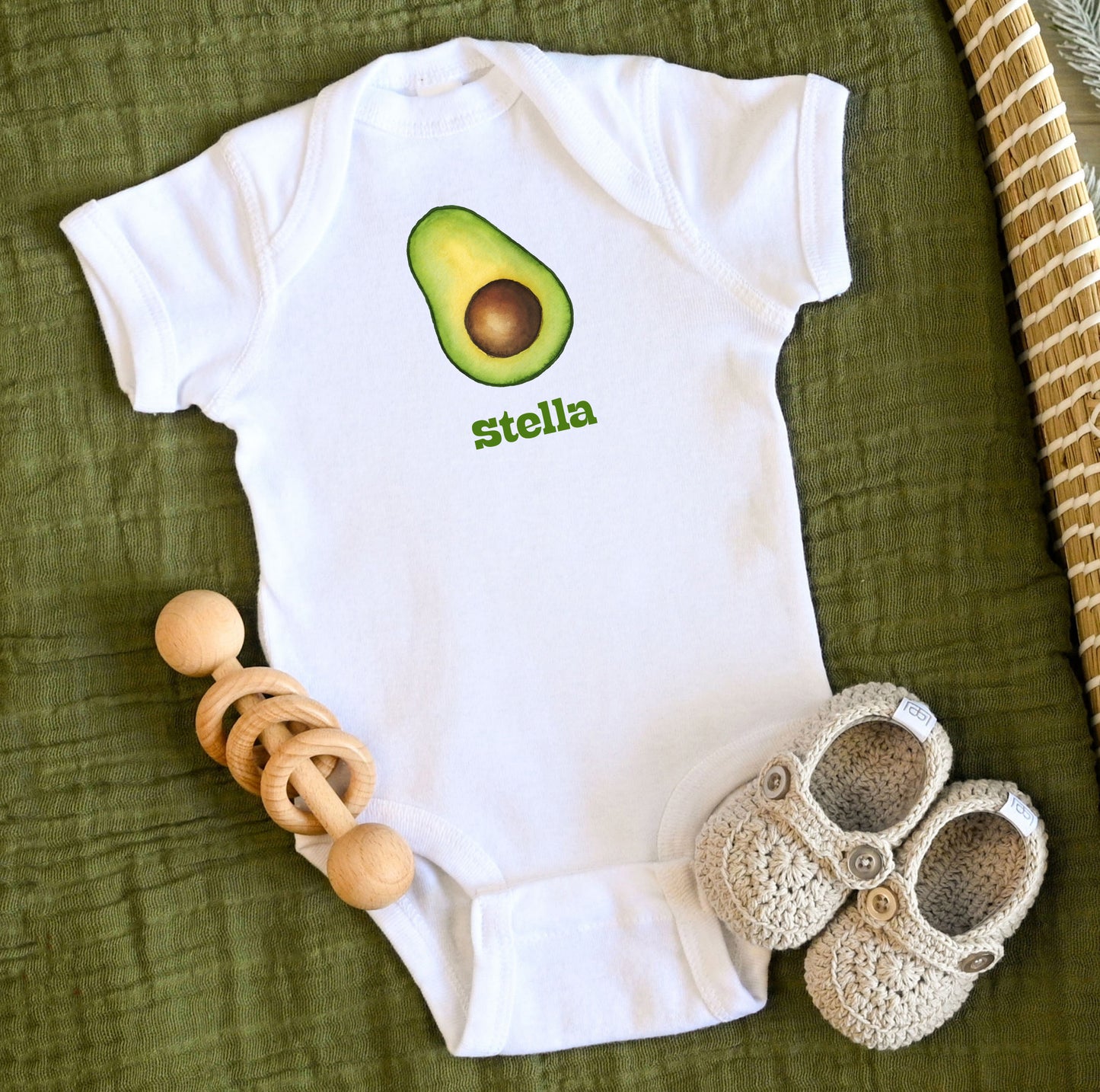 White baby onesie with avocado design and custom personalized text, accompanied by wooden teething ring and crochet shoes on a green fabric background.
