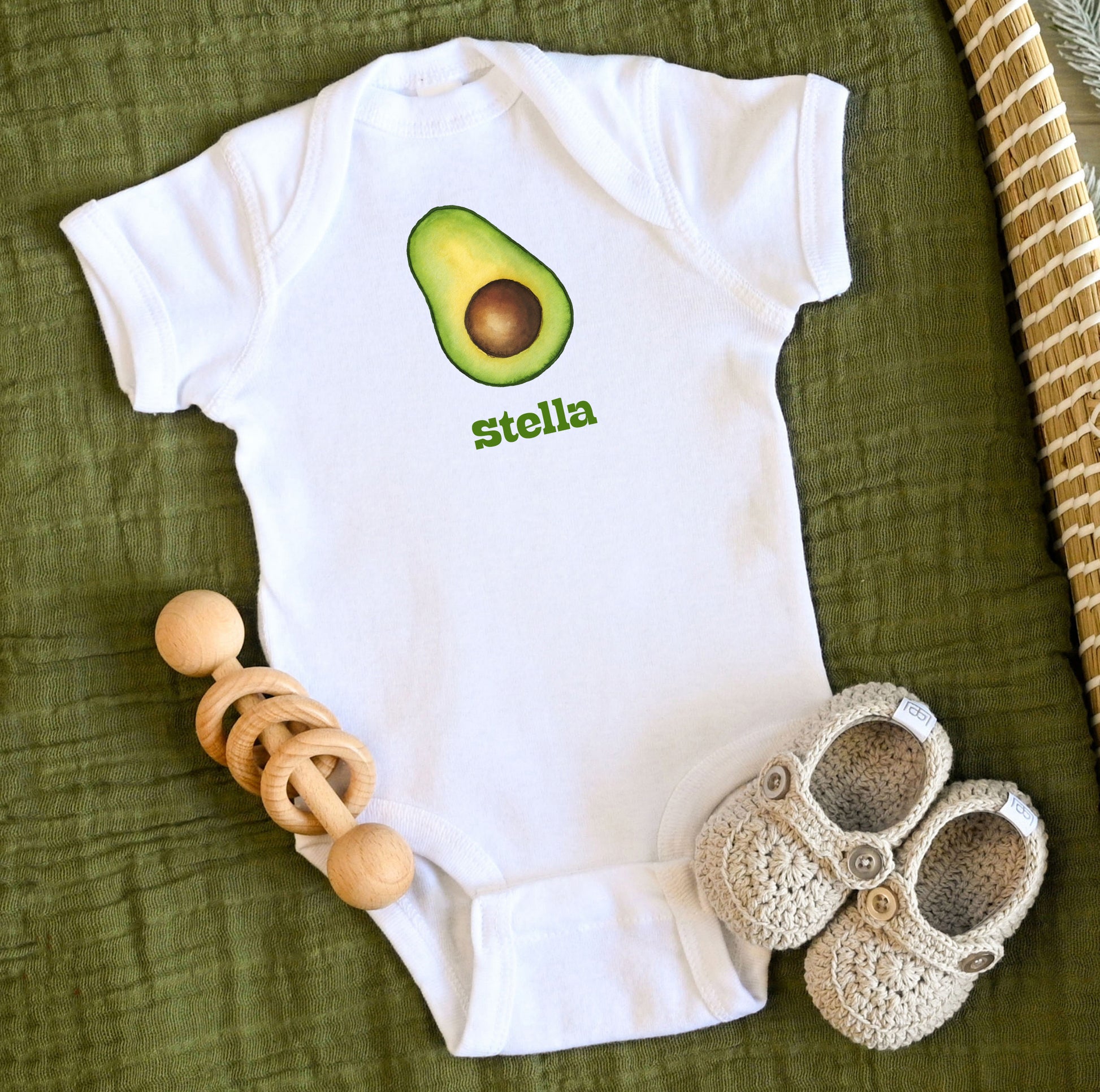 White baby onesie with avocado design and custom personalized text, accompanied by wooden teething ring and crochet shoes on a green fabric background.
