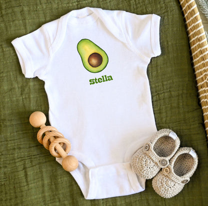 White baby onesie with avocado design and custom personalized text, accompanied by wooden teething ring and crochet shoes on a green fabric background.