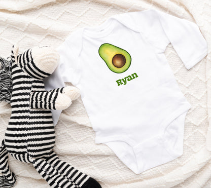 White baby onesie with avocado design and 'Ryan' text, next to a plush toy on a textured surface.