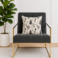 Gray armchair with a decorative warm white pillow featuring black toile pattern, placed in a room with a plant and white walls.
