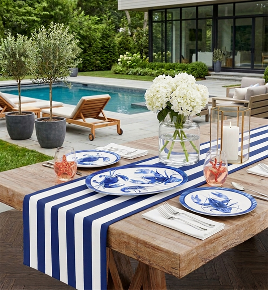 Outdoor dining setup with striped tablecloth, blue and white nautical plates, and white flowers by a pool.