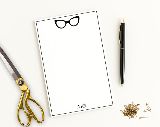Custom notepad paper with a cat eye glasses design, personalized with monogram initials alongside a scissors, pen, and pins on a white background