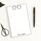 Personalized note pad paper with a name and glasses illustration, surrounded by stationery items on a white background