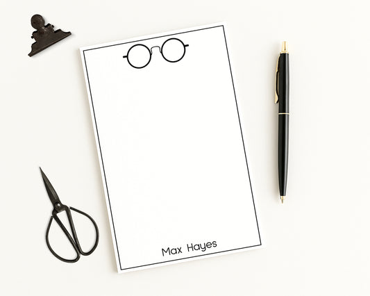 Personalized note pad paper with a name and glasses illustration, surrounded by stationery items on a white background