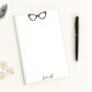 Personalized notepad features cat eye eyeglasses with custom monogram name, and beige border - shown with desk accessories.