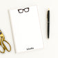 Personalized stationery notepad with chunky tortoiseshell eyeglasses icon and custom name, shown with desk accessories.