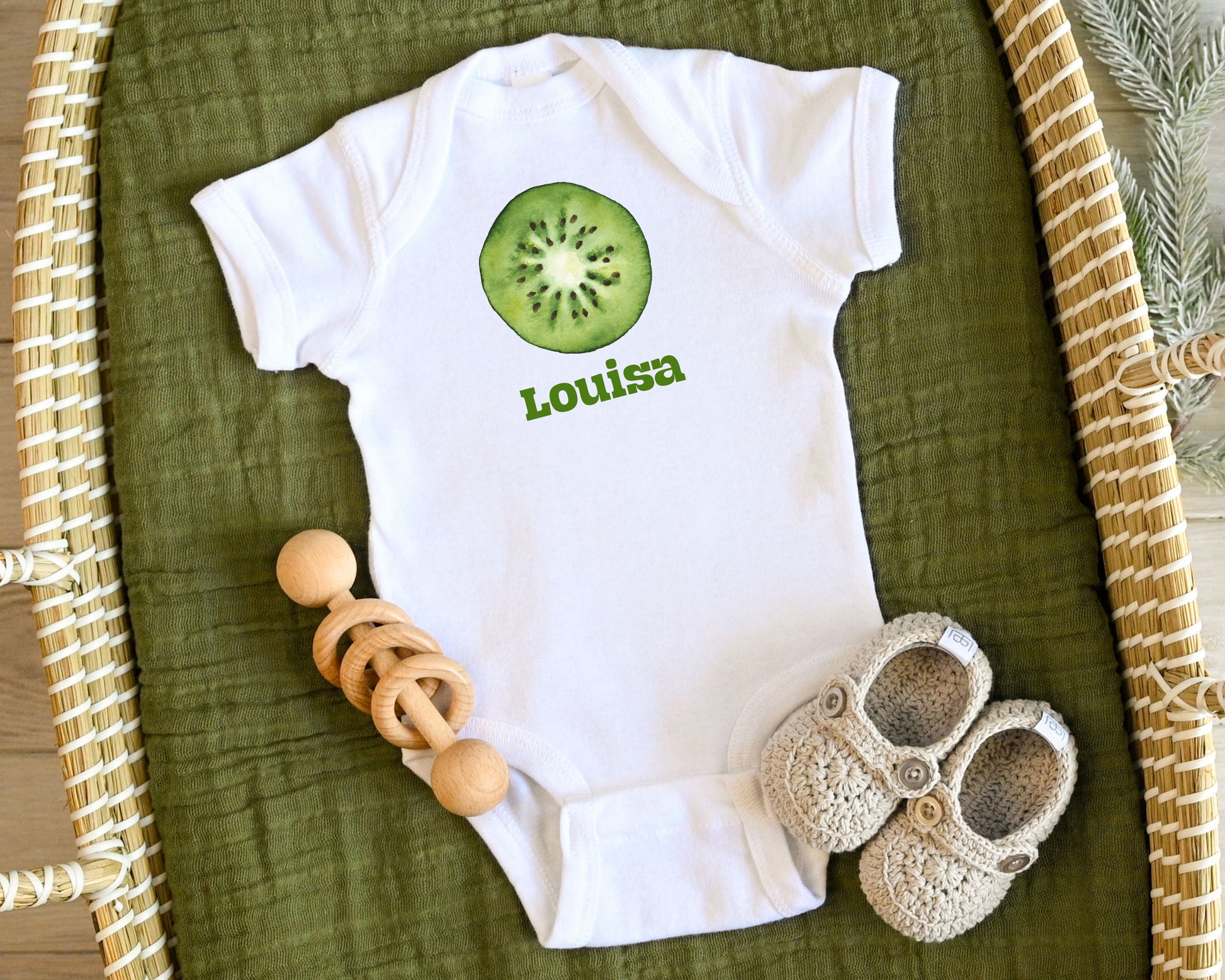 White baby onesie with a kiwi design and name on a green surface with toys.