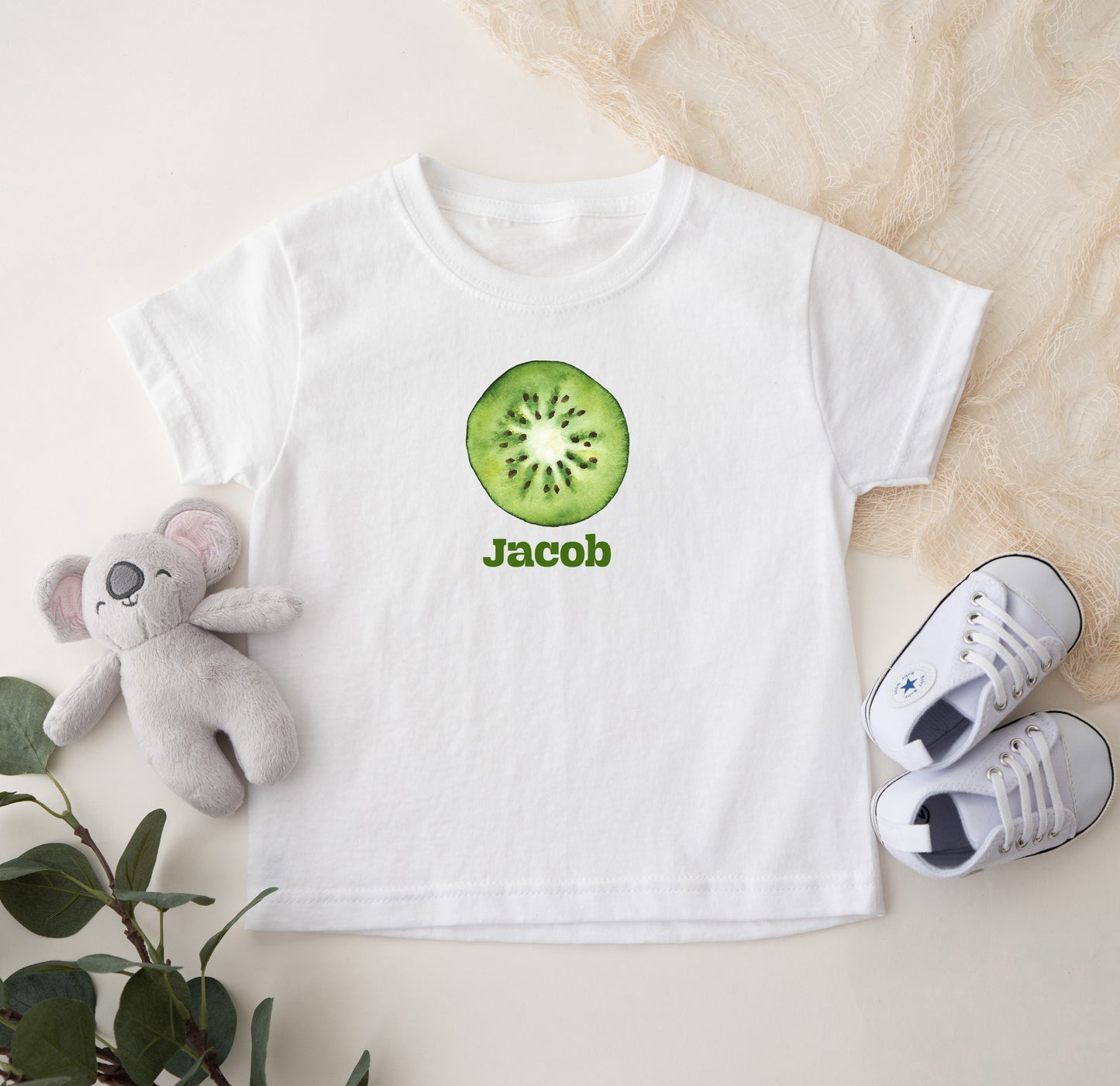 White t-shirt with a kiwi design and 'Jacob' text, accompanied by a plush toy and shoes on a light background.