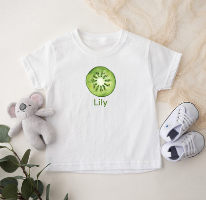 White t-shirt with kiwi design and 'Lily' text, accompanied by a plush toy and shoes on a light background
