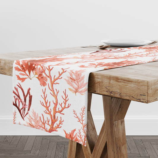 White table runner has red, orange and pink sea coral and seawood pattern. Runner is shown on a wood table.