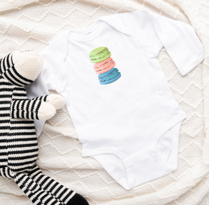 A white long sleeve baby bodysuit with a stacked macaron cookie art print on the front, displayed next to a stuffed animal.