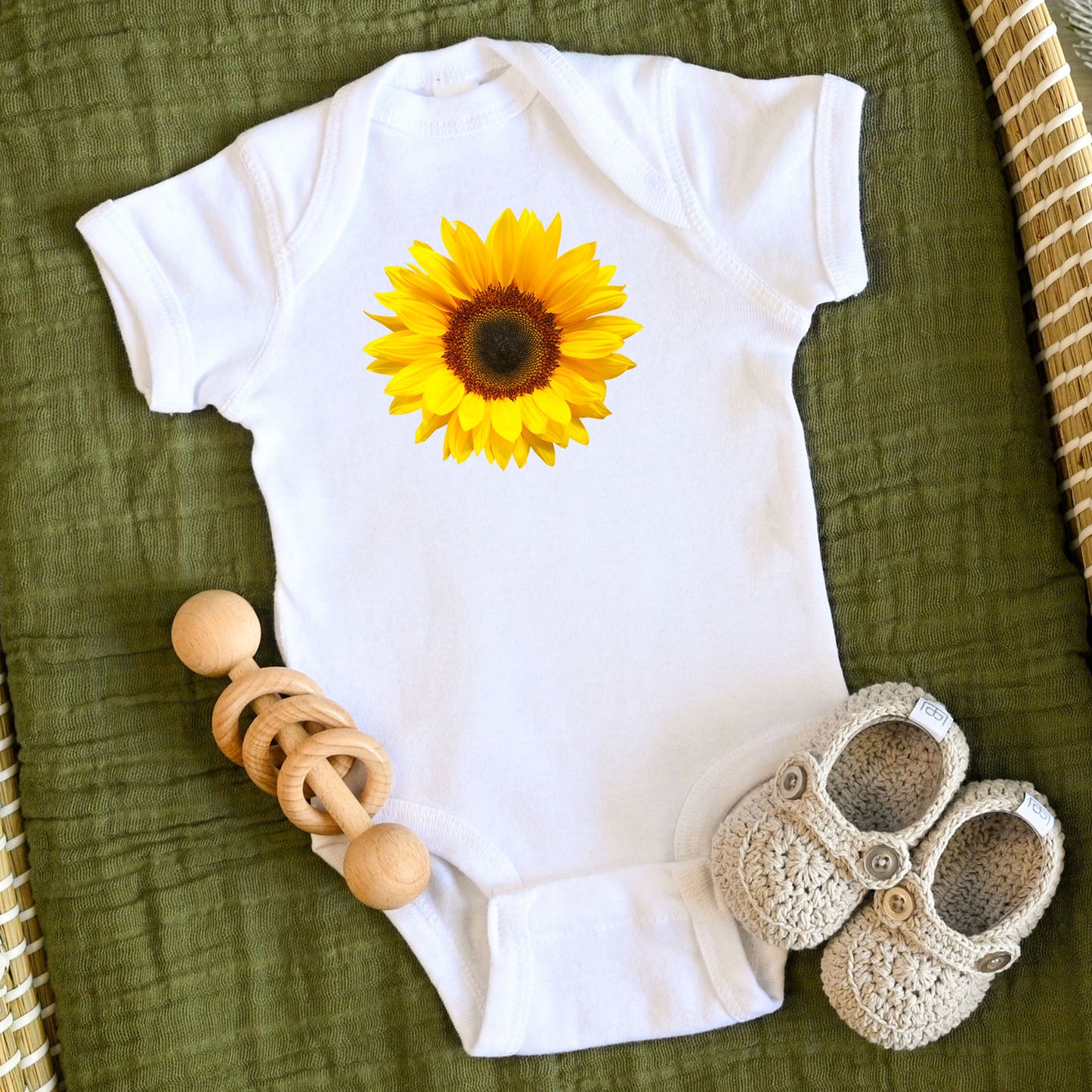 White baby onesie with a sunflower design on a green surface with a wooden rattle and crocheted shoes.