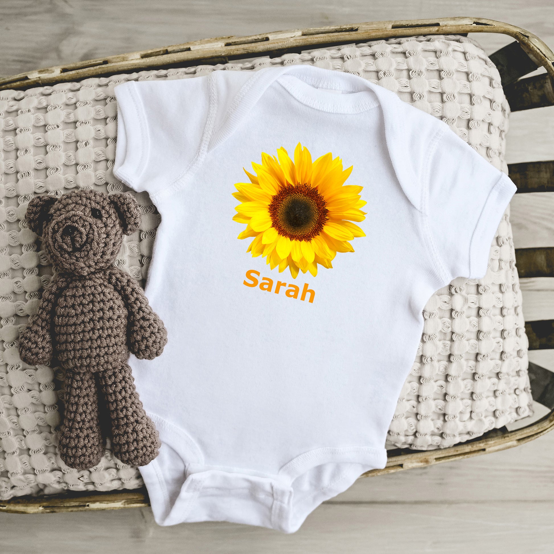 White baby onesie with sunflower design, add any custom text, accompanied by a brown crocheted toy on a woven surface.