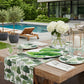 Outdoor dining setup by a pool with tropical tree print table runner and plates, banana leaf platter, coral motif glassware 