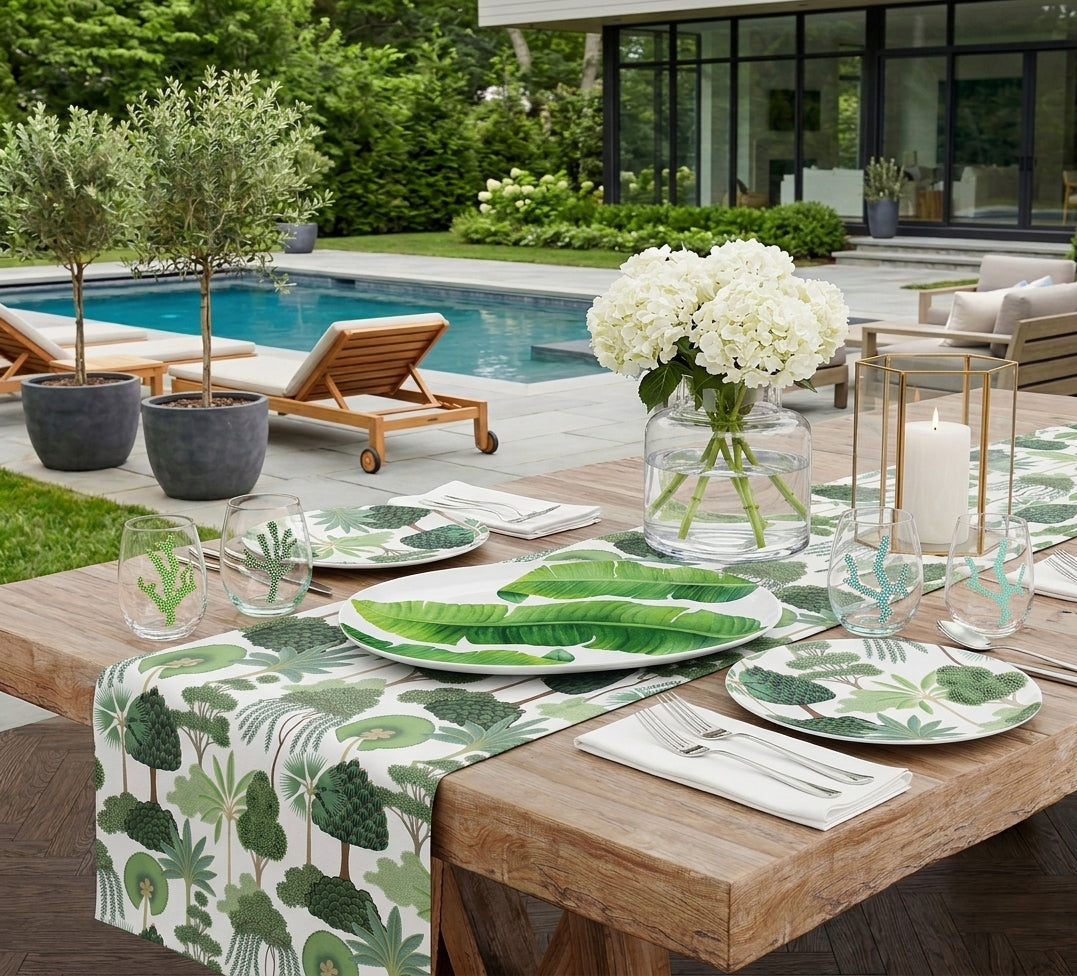 Outdoor dining setup by a pool with tropical tree print table runner and plates, banana leaf platter, coral motif glassware 