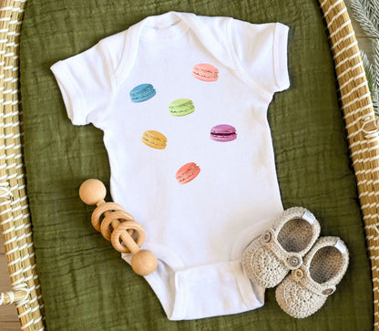 White baby one piece t-shirt with colorful macaron cookies printed on the front, accompanied by a wooden ring and baby shoes.
